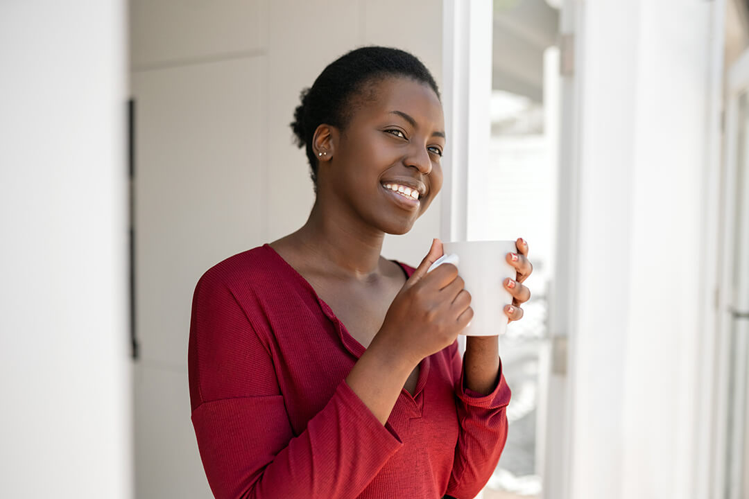 Woman of color in a red sweater holding a white cup of coffee and smiling at the camera.