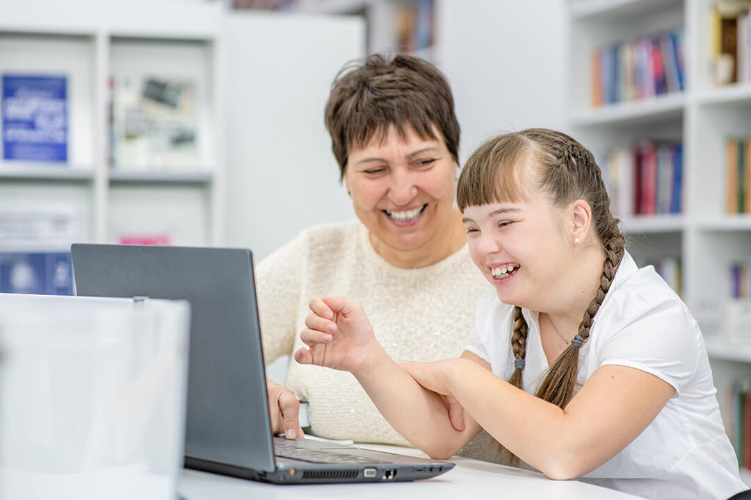 A woman theraphy teacher with an adolescent girl with Downs Syndrome sitting in front of a laptop and smiling.